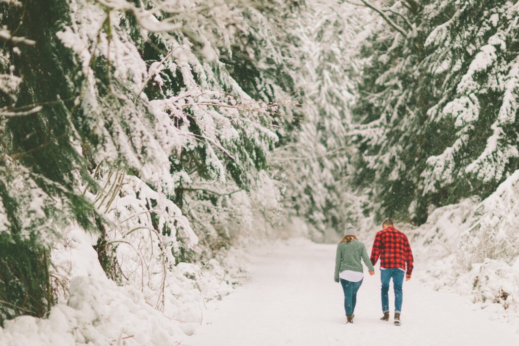 A couple walks hand in hand down a snowy forest path.